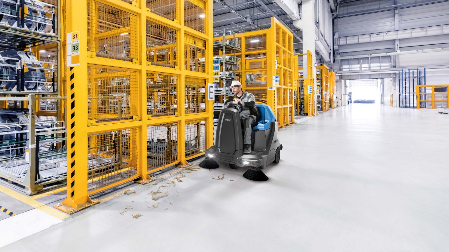 Industrial ride-on sweeper clearing debris beside warehouse storage cages