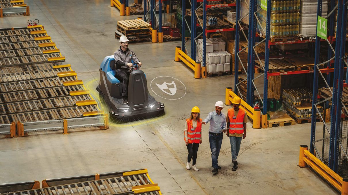 Ride-on sweeper working a distribution centre aisle while staff walk the floor safely